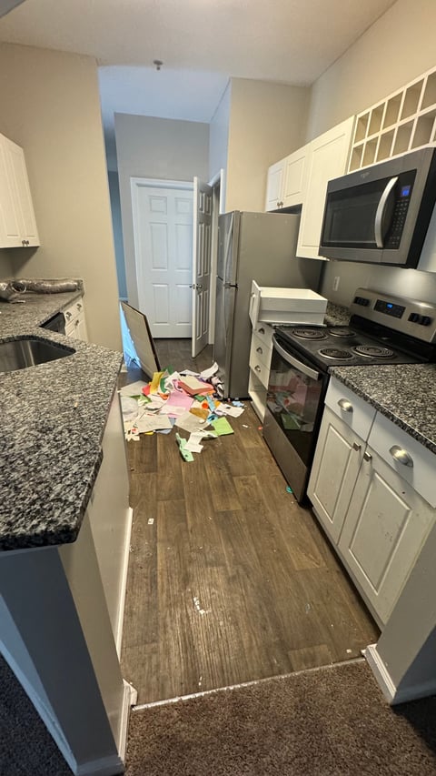 Messy narrow kitchen with white cabinets, stainless steel appliances, and scattered items on the wooden floor