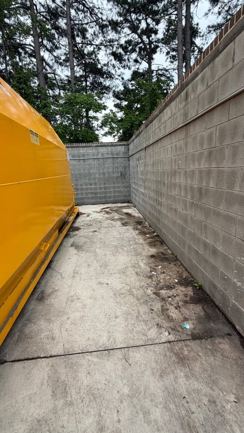Narrow alleyway between a yellow dumpster on the left and gray concrete wall on the right, with trees visible overhead