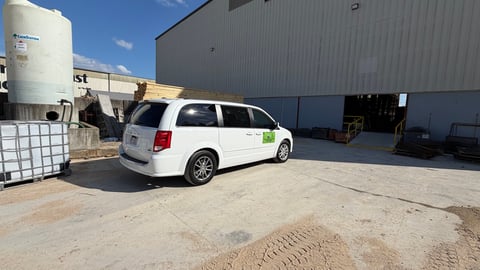 White minivan parked at an industrial facility with a water tank and metal buildings in the background