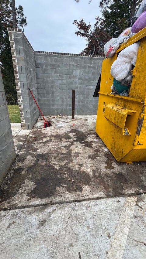 Yellow waste management truck unloading bags into concrete containment area surrounded by trees