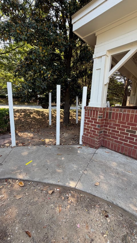 House porch construction with white vertical posts, brick wall, concrete slab, and large trees providing shade