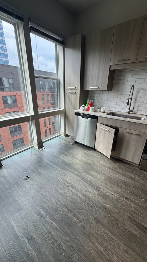 Modern apartment kitchen with wooden cabinets, white subway tile backsplash, stainless steel appliances, and floor-to-ceiling windows overlooking city buildings