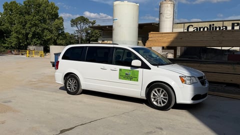 White minivan with green badge parked in industrial lot with large storage tanks and brick building in background
